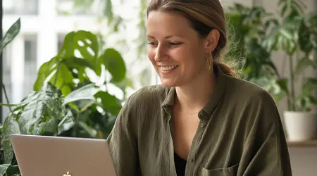 Een vrouw in de 30 die op een laptop werkt met planten in de achtergrond.