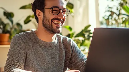 Een man in de 30 met een baard en bril, grijze trui die glimlachend aan een laptop werkt.